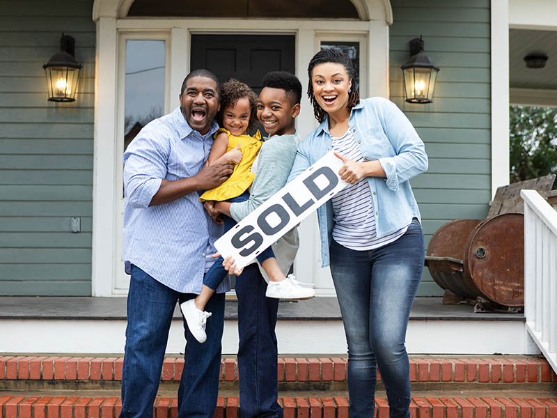 A man, woman, and young boy holding a toddler standing in front of a house holding a sign with "SOLD" printed on it.